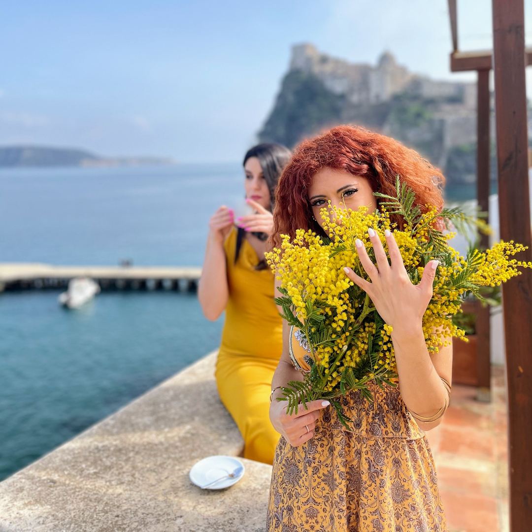 A woman with red hair holds yellow mimosa flowers while overlooking a scenic coastal bay with rocky cliffs and boats in the distance.