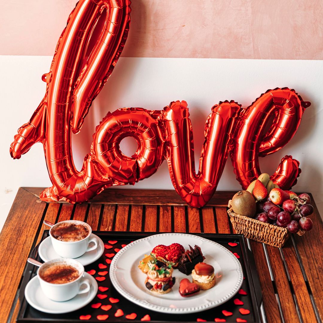 A romantic Valentine's Day breakfast setup featuring red 'Love' balloons, espresso cups, gourmet pastries with fresh berries, and seasonal fruit.