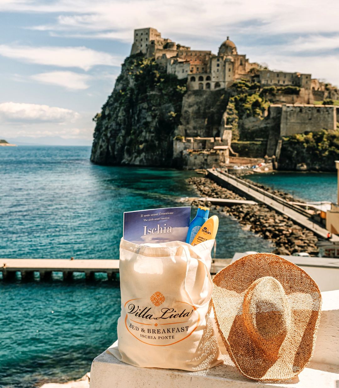 A branded tote bag and straw hat rest on a terrace overlooking the turquoise waters and historic Aragonese Castle of Ischia.