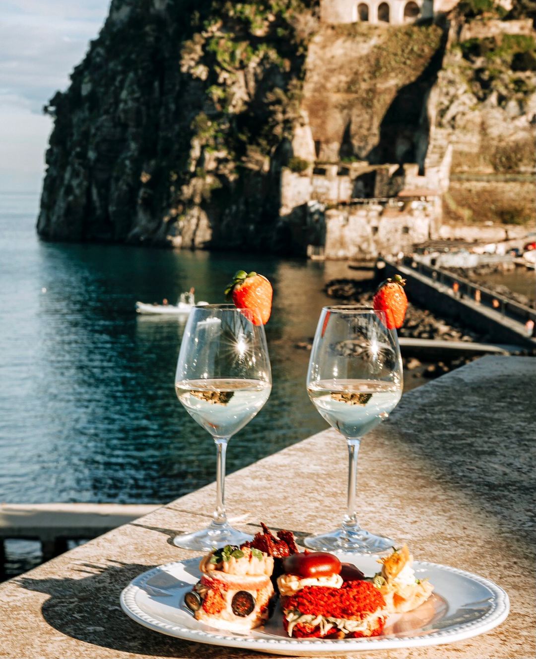 Two glasses of white wine with fresh strawberries and a plate of pastries overlooking a dramatic coastal cove with historic stone architecture.