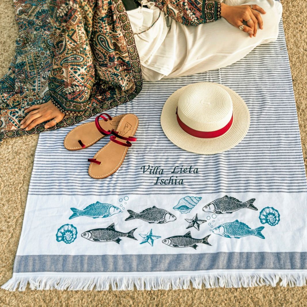 A beach towel branded with Villa Lieta Ischia displays nautical motifs including fish and shells, arranged with sandals and a straw hat on sand.