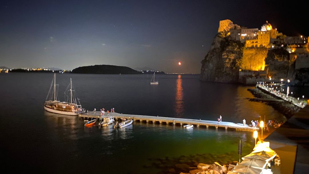 Nighttime view of Aragonese Castle illuminated on the rocky coastline with sailboats moored in the bay below.