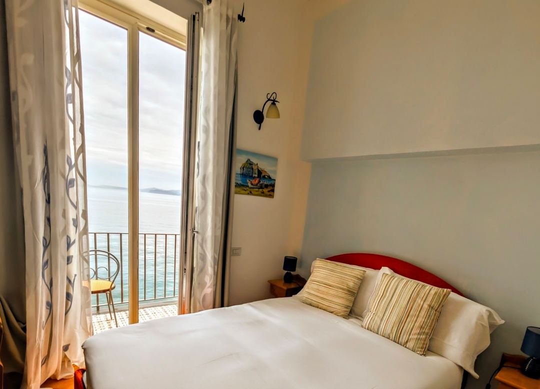 A bright bedroom with a queen bed, striped throw pillows, and sea views through open French doors leading to a waterfront balcony.