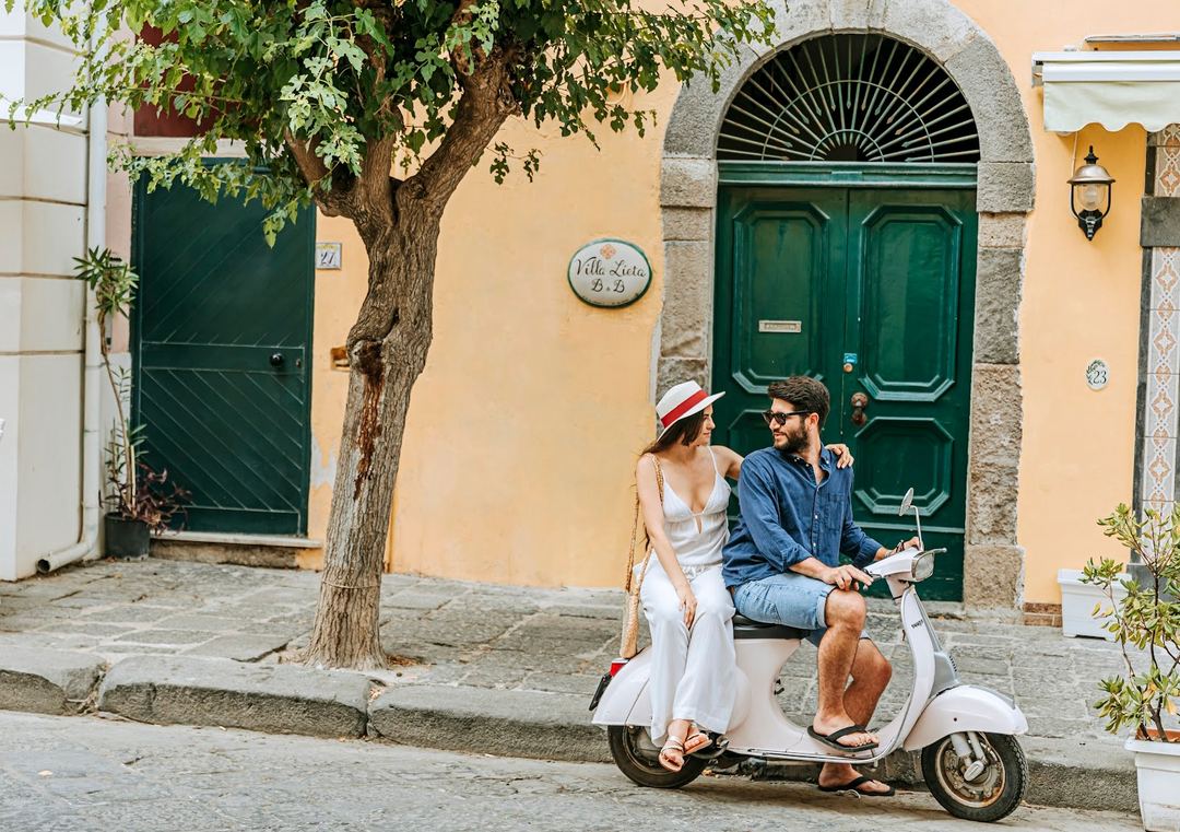 A couple enjoys a Vespa scooter ride in front of Villa Lieta's charming yellow stone building with green arched doors in Ischia.